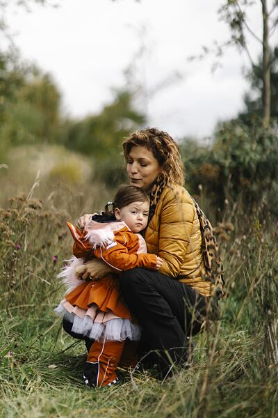 Žena v péřové bundě objímá a uklidňuje malé dítě oblečené v oranžové outdoorové kombinéze Weedo se sukní, stojí spolu v přírodě mezi vysokou trávou a keři. Dětská tutu sukně Weedo ke kombinéze SquirDo