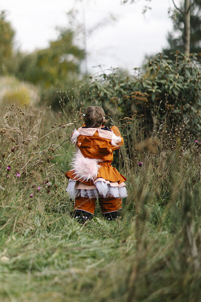 Oranžová dětská outdoorová kombinéza s růžovými a bílými detaily ve tvaru veverky, doplněná o huňatý růžový ocas na zádech, na dítěti stojícím zády v přírodě.