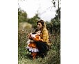 Žena v péřové bundě objímá a uklidňuje malé dítě oblečené v oranžové outdoorové kombinéze Weedo se sukní, stojí spolu v přírodě mezi vysokou trávou a keři. Dětská tutu sukně Weedo ke kombinéze SquirDo